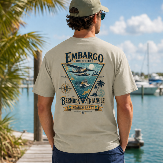 Man wearing a beige t-shirt with 'Embargo Outfitters' design by a marina.