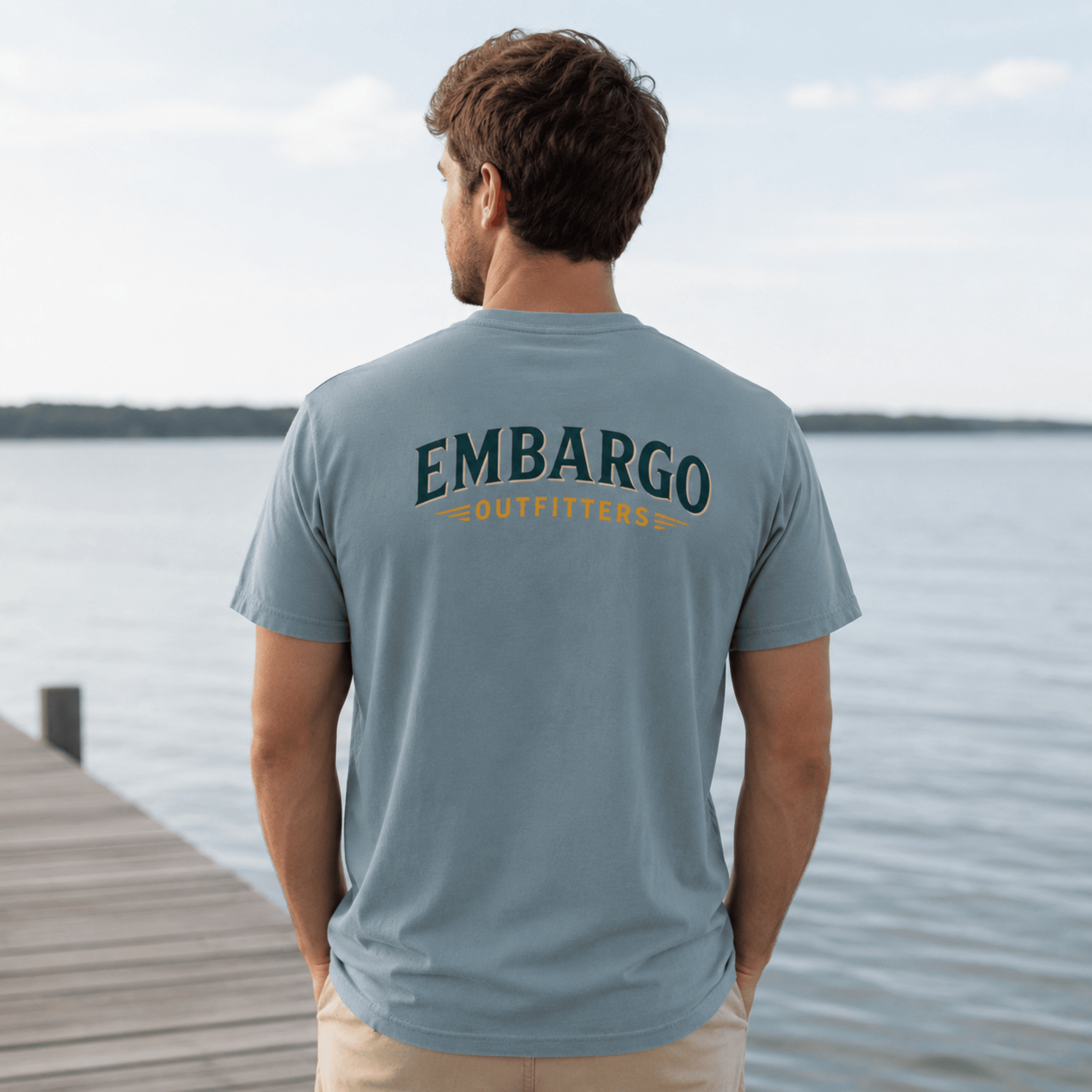Man wearing a gray t-shirt with 'EMBARGO Outfitters' logo by a lake.