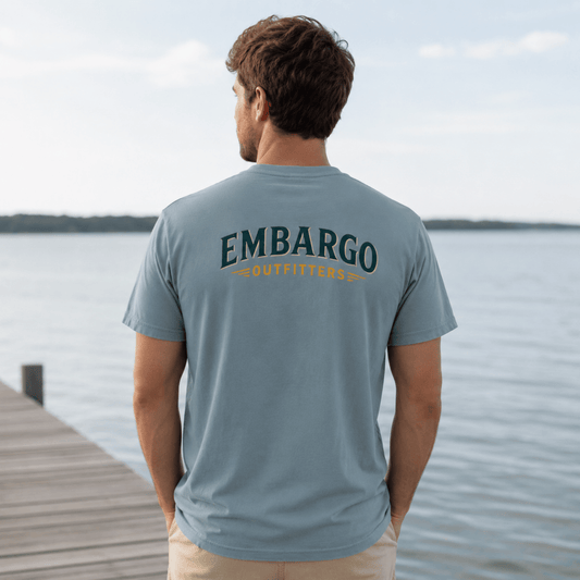 Man wearing a gray t-shirt with 'EMBARGO Outfitters' logo by a lake.