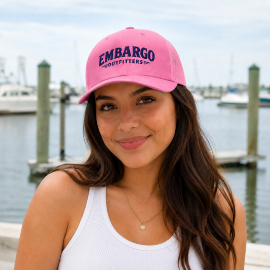 Woman wearing a pink cap with 'Embargo Outfitters' text by a waterfront.