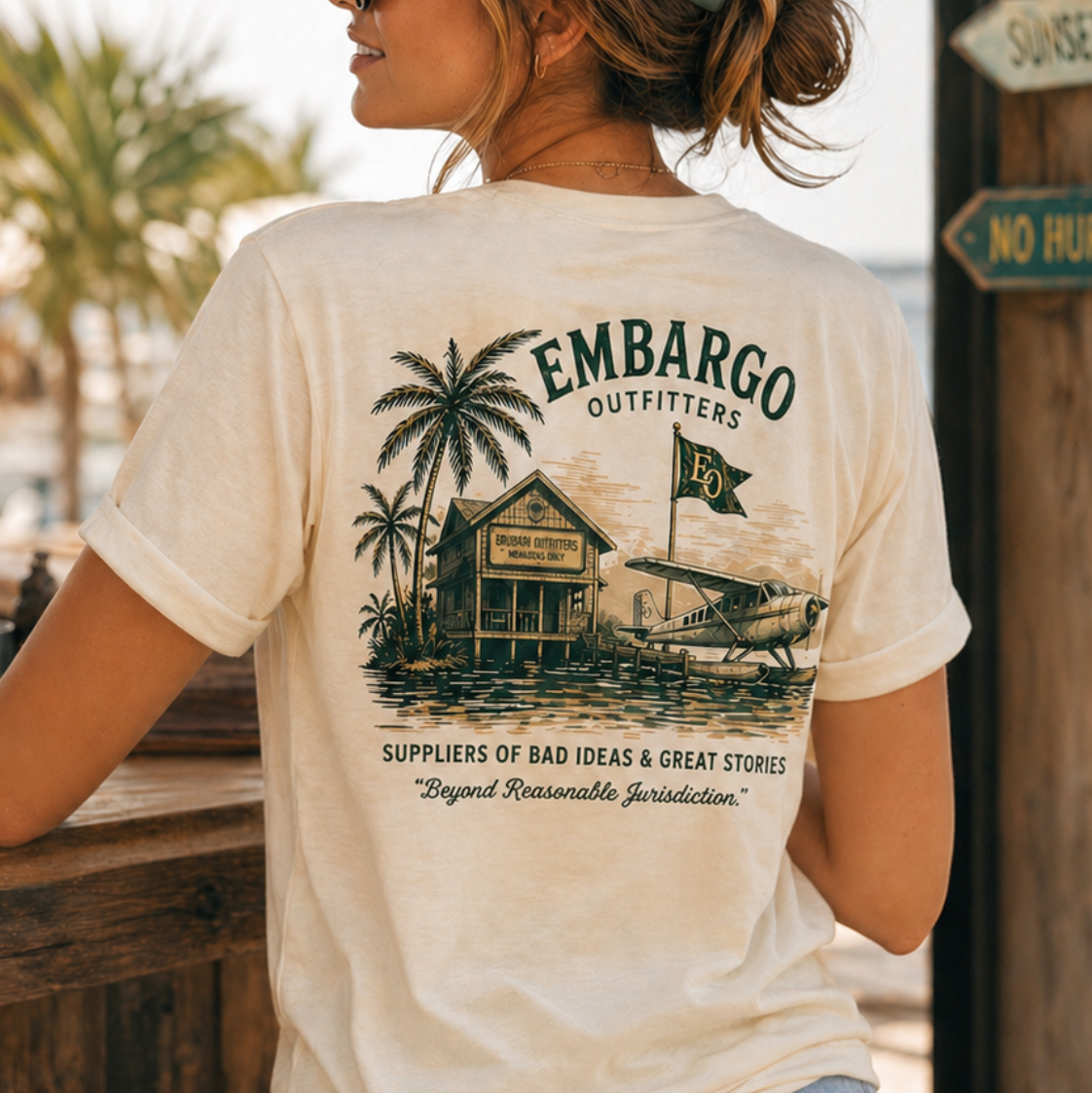 Woman wearing a 'Embargo Outfitters' t-shirt by a dock with palm trees and a signpost in the background.