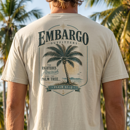 Person wearing a beige t-shirt with a palm tree design and 'Embargo Outfitters' text, standing in front of palm trees.