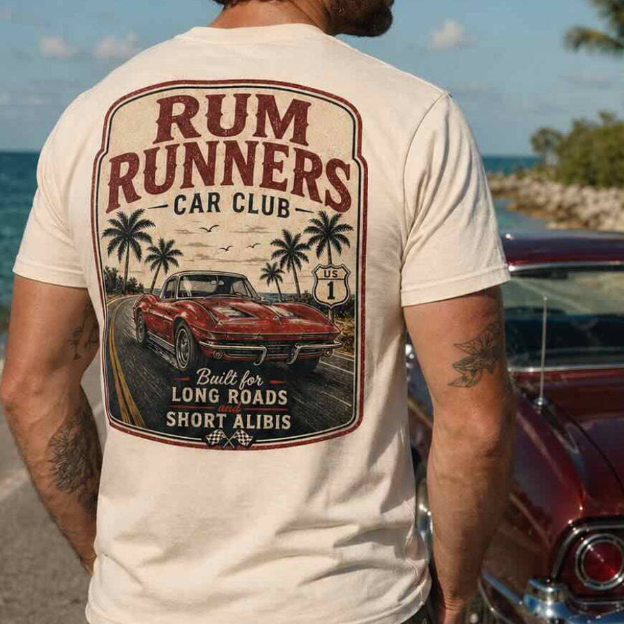 Man wearing a 'Rum Runners Car Club' t-shirt by a vintage car on a scenic road.