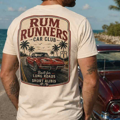 Man wearing a 'Rum Runners Car Club' t-shirt by a vintage car on a scenic road.