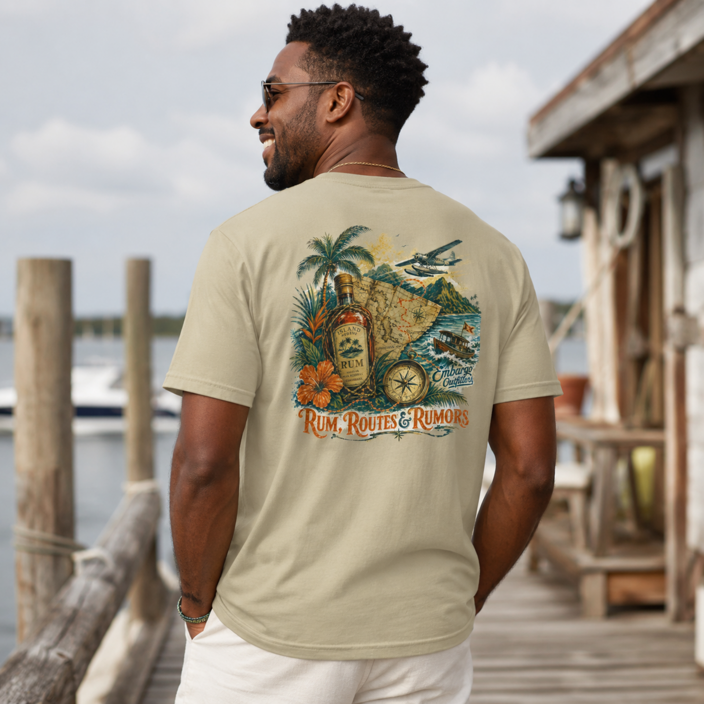 Man wearing a 'Rum, Routes & Rumors' t-shirt by a dock with water and wooden structures in the background.